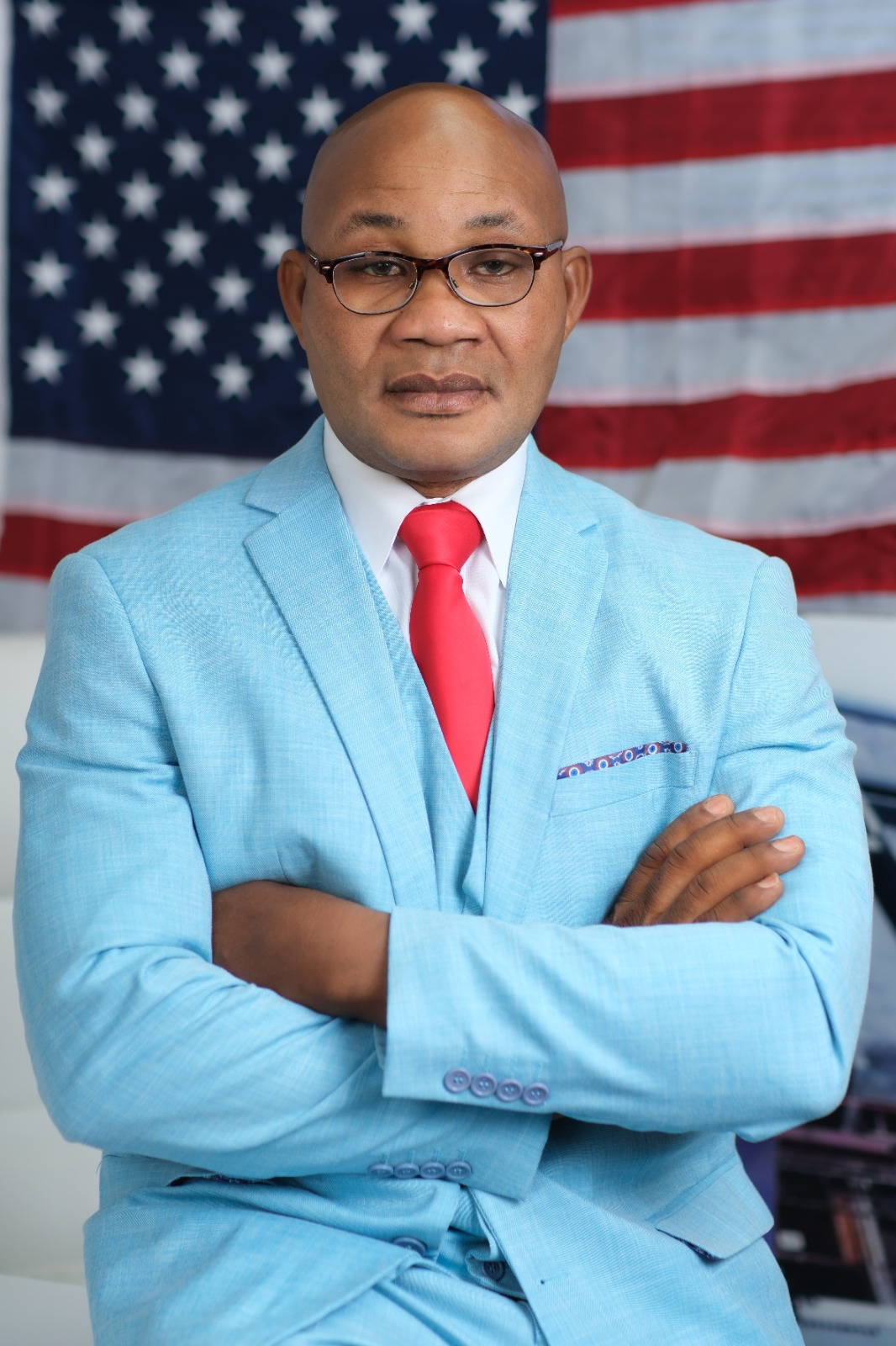 Caneste Succe standing with arms crossed in front of the United States flag